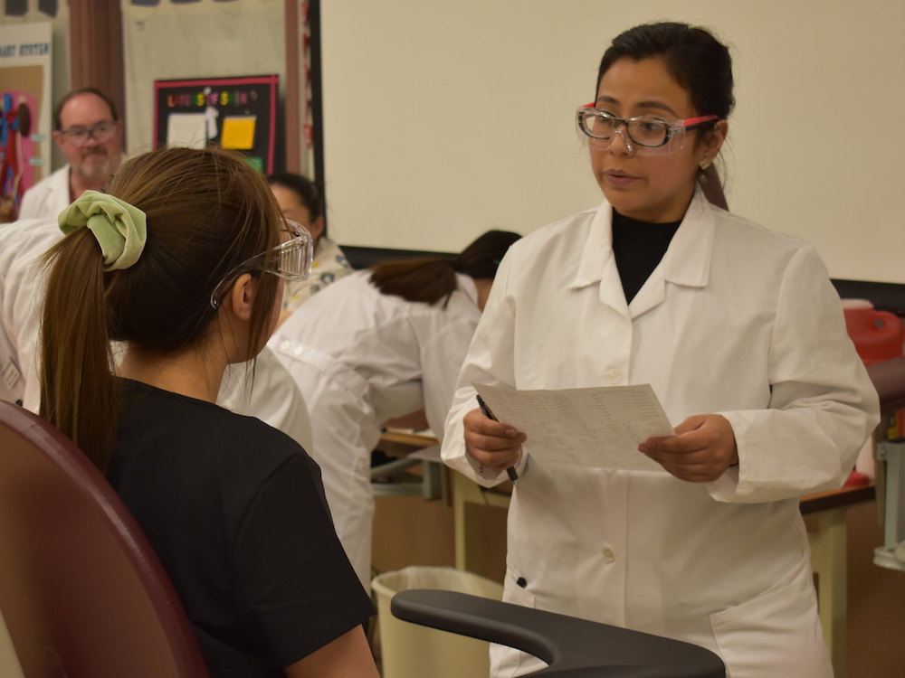 student in white coat talking with seated woman